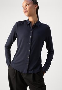 Marineblauwe longsleeve shirt met een kraaghals, knoopsluiting aan de voorkant en een gladde textuur. Gecombineerd met zwarte gestructureerde broek.