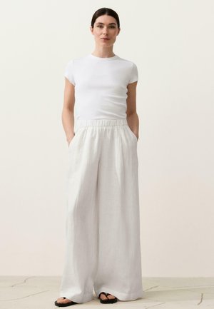 Nő, aki egy testhezálló fehér rövid ujjú felsőt, bőszárú, magas derekú fehér nadrágot és fekete szandált visel, zsebébe dugott kézzel áll.