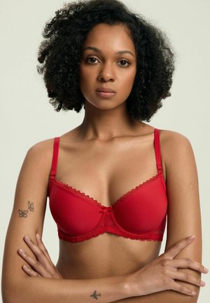 Woman with curly hair wears a red lace bra, arms crossed, with butterfly tattoos on left arm and an airplane tattoo on left wrist.