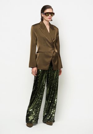 Vrouw draagt een oversized bruine satijnen blazer, grote schildpadzonnebril, donkergroene pailletten wijde broek en bruine schoenen.