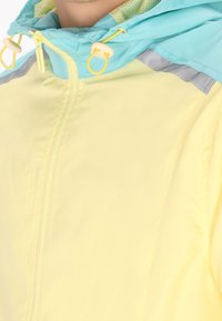 Lichtgele windbreakerjas met een turquoise capuchon, voorzien van grijze reflecterende strepen en trekkoorden op de capuchon voor verstelbaarheid.