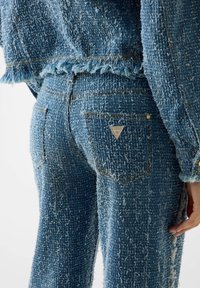 Giacca di denim blu e jeans con una finitura testurizzata, caratterizzati da bordi sfrangiati, cuciture dorate e una targhetta triangolare con il marchio sulla tasca.