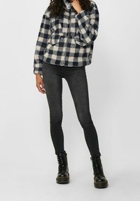 Veste zippée en polaire à motif à carreaux bleu marine et crème, avec deux poches avant et un col montant, associée à un jean skinny gris et des bottes noires.