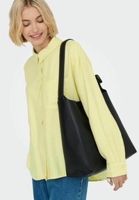 Schwarze Leder-Tote mit glatter Textur und breiten Riemen. Leichtes gelbes Button-up-Hemd mit einer Brusttasche, kombiniert mit blauen Jeans.