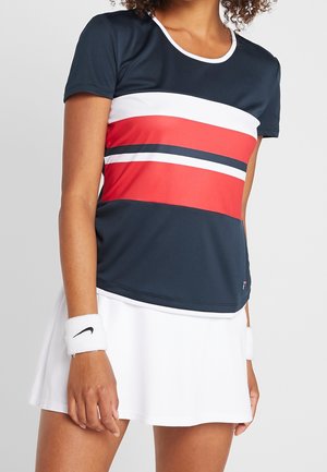 Kobieta w sportowej koszulce w granatowe, białe i czerwone paski, białej spódnicy tenisowej i białych opaskach Nike na obu nadgarstkach.