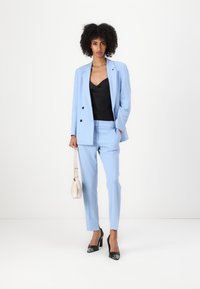 Light blue suit with tailored jacket and slim-fit pants, paired with a black satin camisole and black high-heeled shoes, holding a light bag.