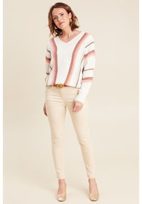 Weißer Strickpullover mit rosa und braunen Streifen, V-Ausschnitt-Design, kombiniert mit hellbeigen, enganliegenden Hosen und Schuhen mit goldenen Akzenten.