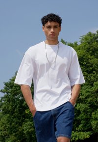 Tricou alb supradimensionat cu mâneci scurte, asortat cu pantaloni scurți din denim albastru. Modelul poartă o colier din lanț argintiu și stă în aer liber, înconjurat de verdeață.