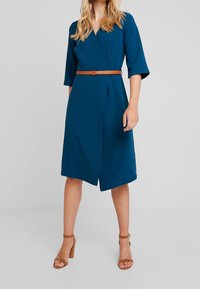 Kvinna iförd en knälång teal omslagsklänning med 3/4 ärmar, ett beige bälte och beige högklackade sandaler, stående mot en neutral bakgrund.