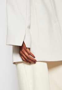 Bílý blejzr s volnějším střihem, s výraznými rameny a ostrým klopou. Ruka spočívá na krémovém oděvu pod ním.