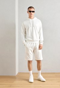 Felpa bianca testurizzata con cappuccio e cordini, abbinata a pantaloni corti coordinati. Indossata con sneakers bianche e occhiali da sole, su un pavimento in legno chiaro.