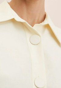 Chemise de couleur crème avec col, dotée de deux grands boutons ronds, texture lisse et design minimaliste.
