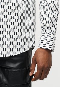 Κοντινό πλάνο του χεριού ενός ατόμου δίπλα σε λευκό πουκάμισο με μαύρο γεωμετρικό σχέδιο και μαύρο δερμάτινο παντελόνι.