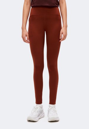 Personne portant des leggings taille haute couleur rouille avec une poche latérale et des chaussures de sport blanches, debout devant un fond uni.