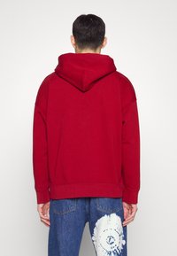 Czerwony sweter z kapturem, z luznym krojem i ściągaczami. Noszony z niebieskimi dżinsami, które mają wzór tie-dye na jednej nodze.