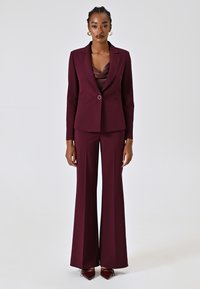 Bordeaux pak met een getailleerde blazer en brede broekspijpen. Satijnen top met een kraag, gladde textuur, zilveren knoopdetail op de blazer.