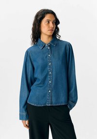 Denimskjorta i medelblått, med lång ärm, knappstängning fram och klassisk krage. Mjuk textur med en något avslappnad passform.