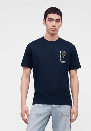 Giovane uomo che indossa una t-shirt nera Lacoste con una piccola grafica di racchetta da tennis e testo sul petto sinistro, abbinata a jeans azzurri.