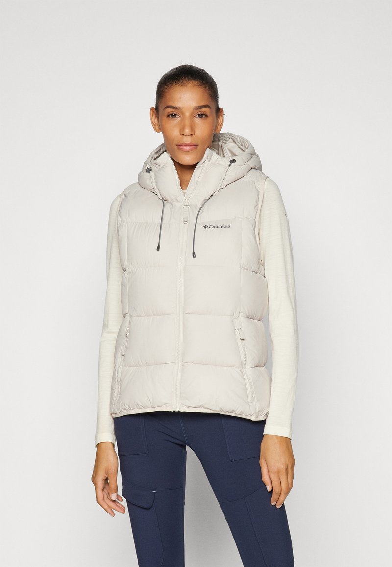 Vrouw in een lichtbeige gewatteerde vest met capuchon, gedragen over een lange mouwtop en marineblauwe cargo broek, staande tegen een effen achtergrond.
