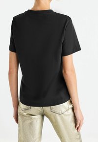 Sort bomulds t-shirt med korte ærmer og rund hals, vist fra ryggen. Båret med metalisk guld bukser, der har en glat tekstur.