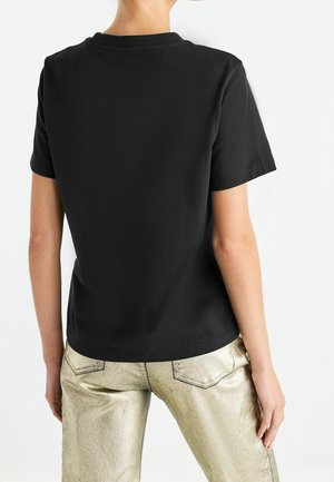 Sort bomulds t-shirt med korte ærmer og rund hals, vist fra ryggen. Båret med metalisk guld bukser, der har en glat tekstur.