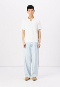 Camisa de polo blanca de punto con mangas cortas, combinada con vaqueros de pierna ancha azul claro y zapatos de lona beige, sobre un fondo liso.