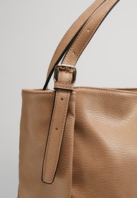 Borsa tote in pelle beige con superficie texture. Presenta una lunga tracolla fissata da una fibbia dorata e impunture nere.