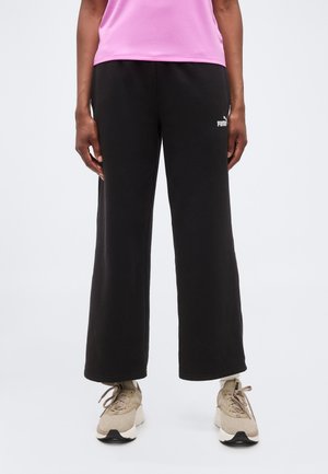 Osoba nosiaca čierne široké Puma tepláky, béžové tenisky a ružový top, stojaca na bielom jednofarebnom pozadí.