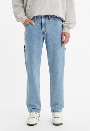Pantalones vaqueros de denim azul con corte relajado y bolsillos laterales, combinados con un sudadera gris claro y zapatillas blancas. Manos parcialmente en los bolsillos.