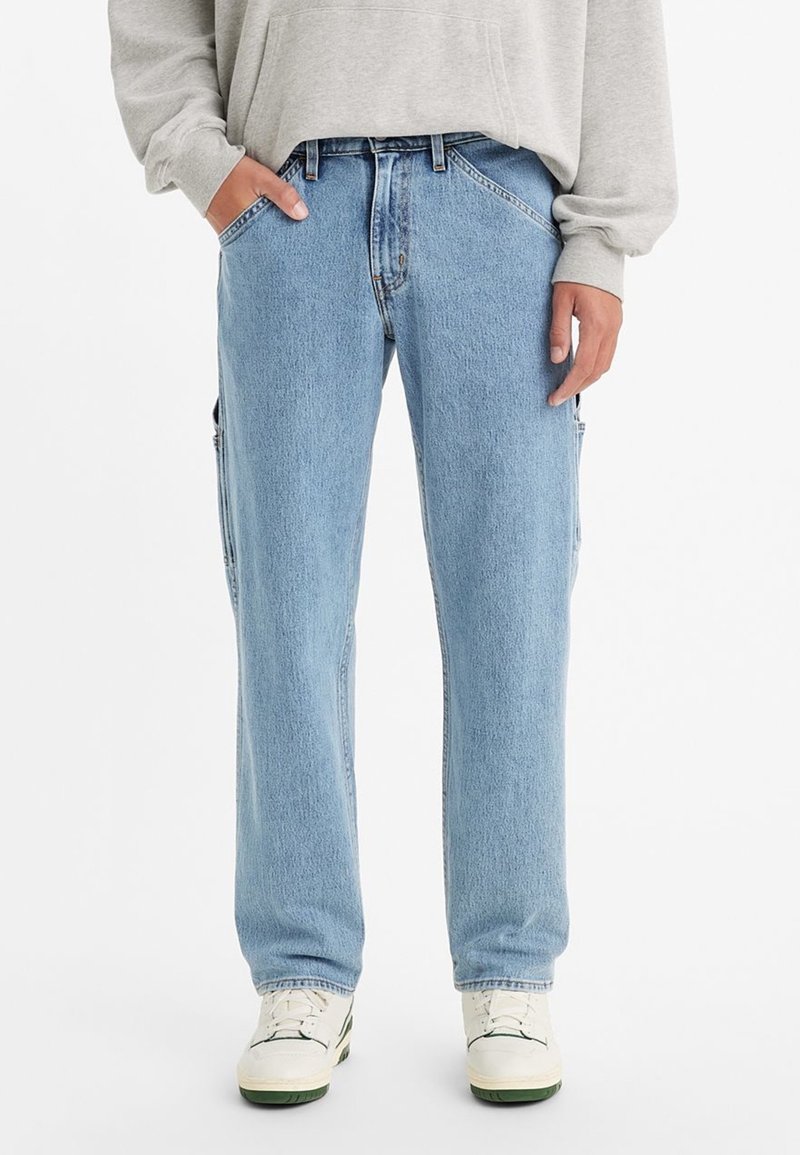 Blå denimjeans med avslappnad passform och sidofickor, bärs med en ljusgrå sweatshirt och vita sneakers. Händerna delvis i fickorna.
