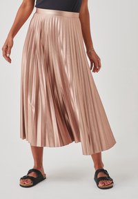Plissé midi rok in zacht roze satijn, met een hoge tailleband, glanzende afwerking en een vloeiende silhoutte. Gecombineerd met zwarte sandalen.