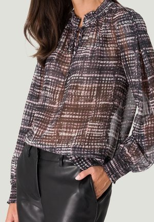 Femme portant un chemisier transparent à manches longues avec motif, col et poignets à volants, associé à un pantalon en cuir noir, main dans la poche.