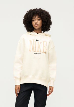 Jeune femme aux cheveux bouclés portant un sweat à capuche Nike crème oversize avec des lettres beiges et un pantalon noir, debout devant un fond uni.