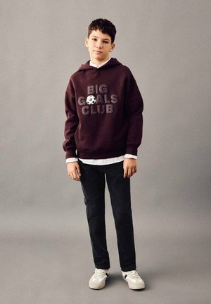 Fant z majico s kapuco bordo barve z napisom "BIG GOALS CLUB" in grafiko nogometne žoge, črnimi hlačami in belimi supergami, stoji na enobarvnem ozadju.