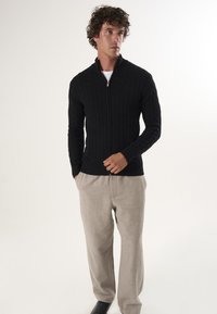 Maglione nero in maglia con zip e motivo a trecce, abbinato a pantaloni beige chiaro e scarpe nere, esposto in un ambiente neutro.