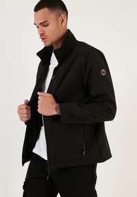Chaqueta softshell negra con un alto cuello, bolsillos con cremallera y un sutil parche de logo. Presenta una textura suave y una forma ajustada.