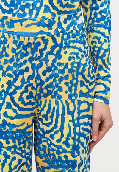 Κίτρινο και μπλε τοπ μακρυμάνικο με αφηρημένο σχέδιο και παντελόνι που ταιριάζει, κοντινή λήψη στον κορμό και το δεξί χέρι στο πλάι.