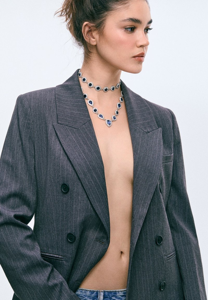 Femme aux cheveux attachés porte des colliers superposés de pierres précieuses bleues et un blazer oversize gris foncé à fines rayures, laissant apparaître la poitrine nue sous la veste.