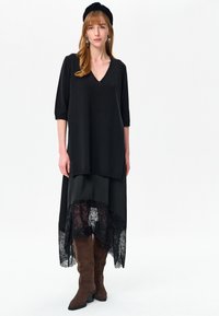 Kobieta ubrana w czarną tunikę z dekoltem w serek, przezroczystą czarną spódnicę z haftem, czarną opaskę na głowę, zwisające kolczyki oraz brązowe zamszowe kozaki do kolan.