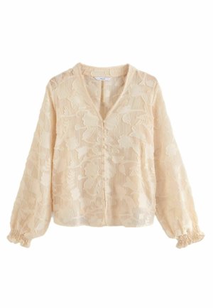 Kobieta w beżowej, teksturowanej bluzce z bufkami na rękawach, wysokim stanem i szerokimi nogawkami w ciemnoniebieskich dżinsach oraz brązowymi zamszowymi butami w szpic.