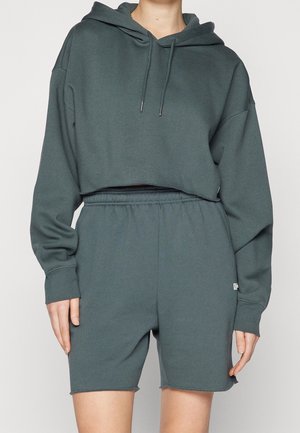 Draagt een donkergroene cropped hoodie met trekkoorden en bijpassende high-waist elastische shorts, zichtbaar van nek tot midden van de dij.