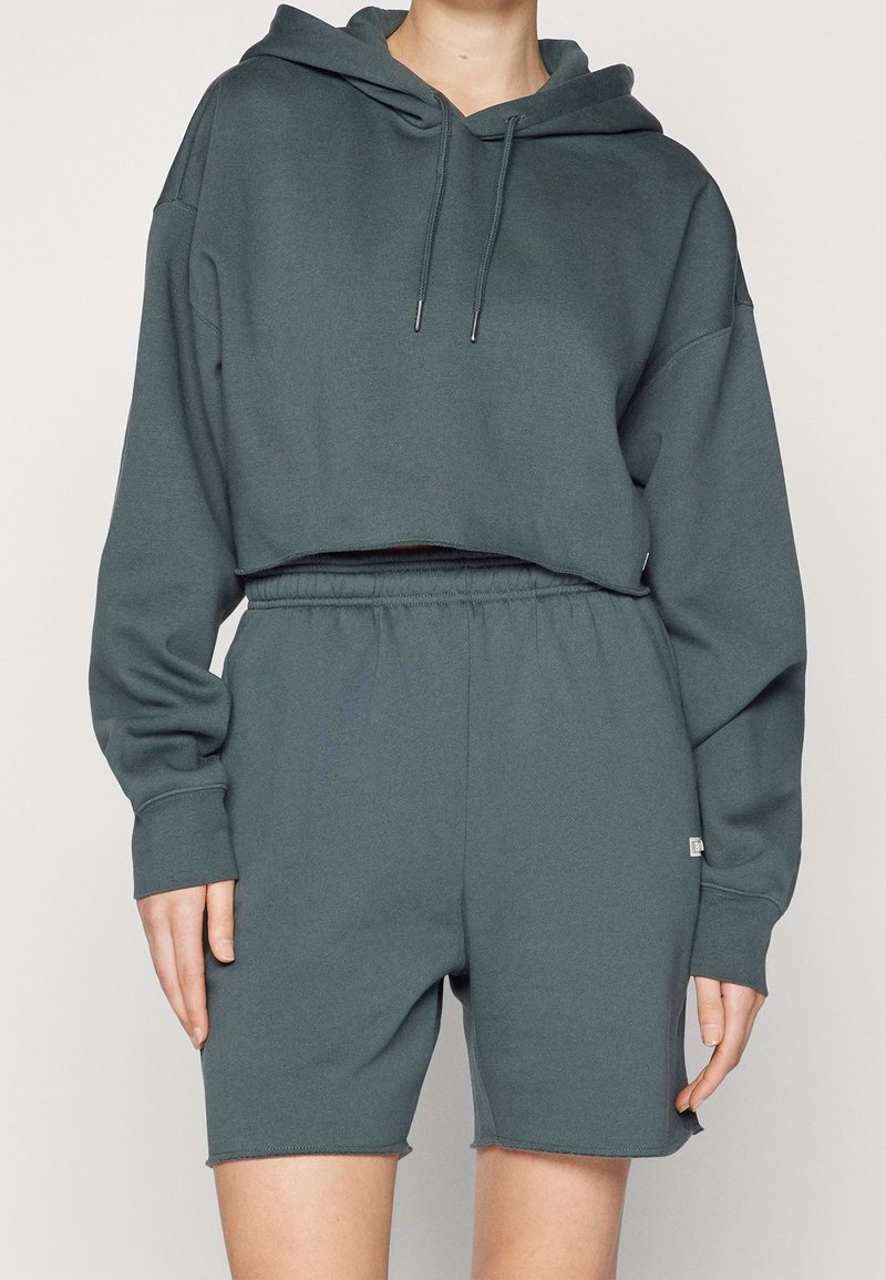 Portant un sweat à capuche court vert foncé avec cordons et un short assorti taille haute avec élastique, visible du cou au milieu des cuisses.