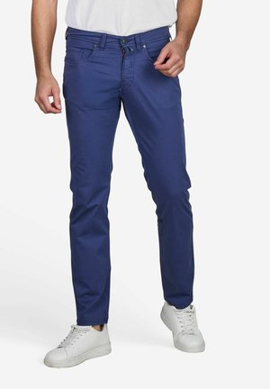 Blue cotton chinos with a slim fit, featuring five pockets, a button fly, and subtle stitching details. Paired with white sneakers.