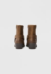 Botas de tobillo de cuero marrón con un acabado suave, cremalleras laterales y una correa decorativa con acentos de latón. Tacón bloque para mayor estabilidad.