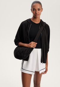 Fekete steppelt válltáska puha, texturált felülettel, keresztben viselve, mellette egy fekete cipzáras kapucnis felső és fehér pliszírozott rövidnadrág.