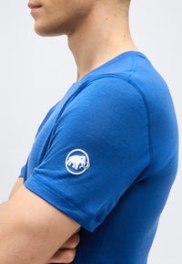 Πλάγια όψη ενός ατόμου που φοράει εφαρμοστό μπλε κοντομάνικο πουκάμισο με λευκό λογότυπο μαμούθ στο αριστερό μανίκι.