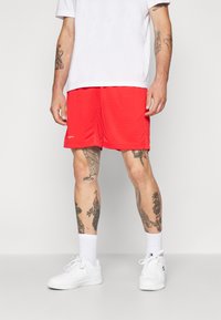 Κόκκινες αθλητικές βερμούδες με δικτυωτή υφή, με λογότυπο στο τελείωμα. Συνδυασμένες με λευκά αθλητικά παπούτσια και λευκές κάλτσες μέχρι τον αστράγαλο.