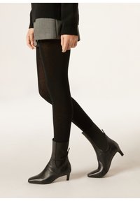 Černé kožené kotníkové boty s špičatou špičkou a nízkým podpatkem, se zipem na boku a hladkým povrchem. Kombinují se s černými punčochami a vrstvenou sukní.