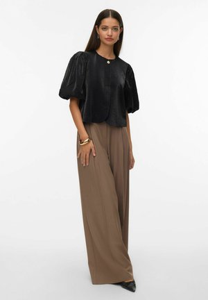 Femme aux longs cheveux foncés portant un haut noir à manches bouffantes avec un bouton unique, un pantalon large marron, un bracelet en or et des chaussures noires à bout pointu, debout.