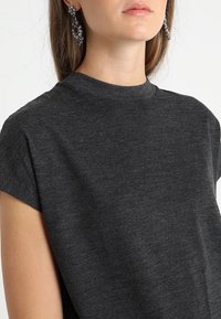 Tmavě šedé tričko s krátkým rukávem a kulatým výstřihem. Měkká, strukturovaná látka s mírným leskem. Náušnice modelky jsou zdobené a třpytivé.